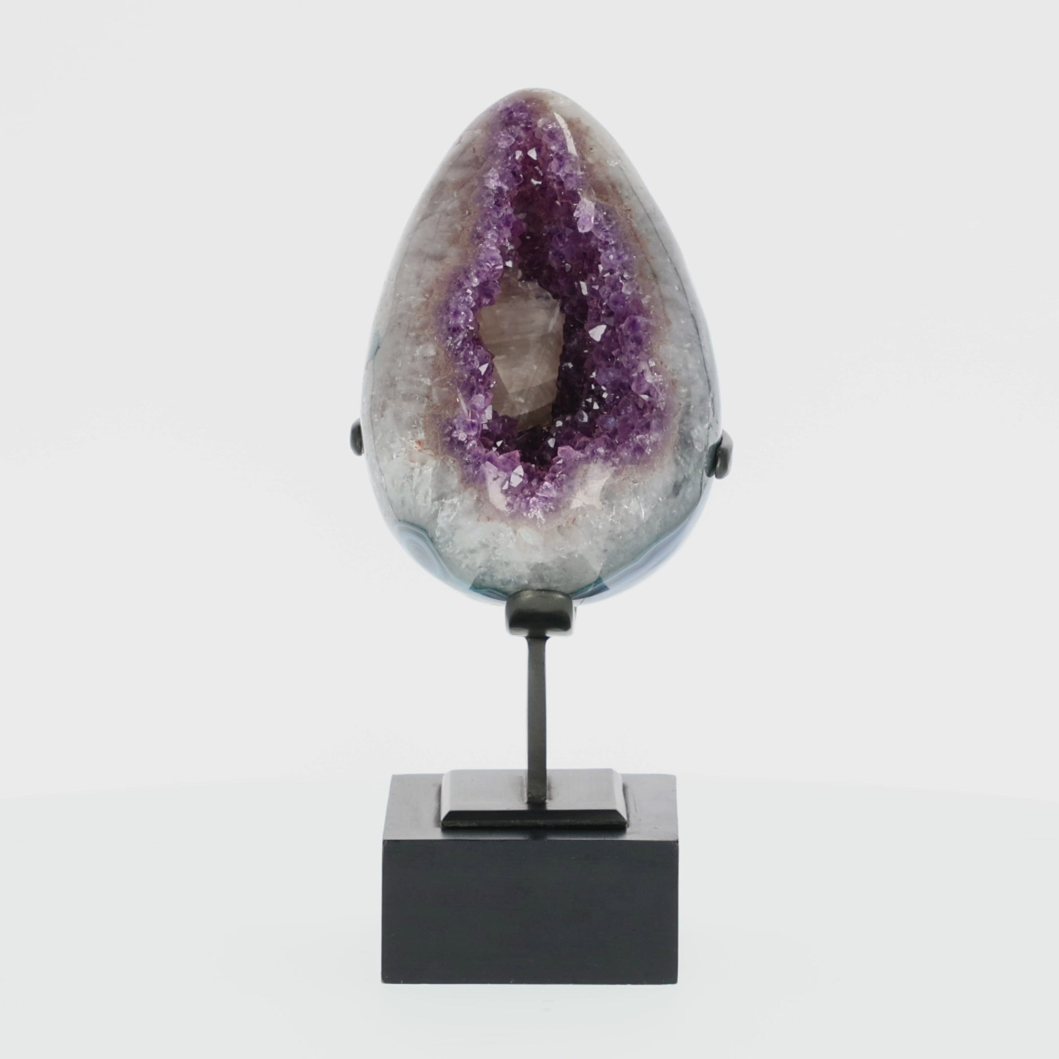 Video laden: Poliertes Amethyst-Ei aus Uruguay auf Metallständer mit sichtbarer Achatstruktur außen und funkelnder Amethyst-Druse im Inneren.