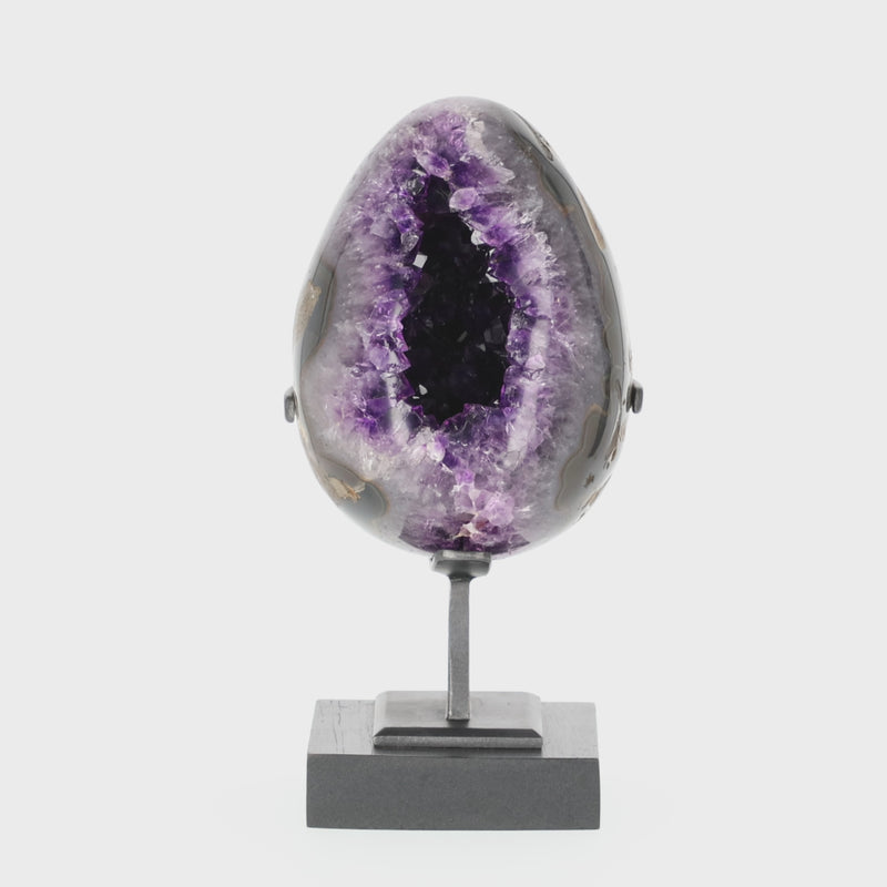 Poliertes Amethyst-Ei aus Uruguay auf Metallständer mit sichtbarer Achatstruktur außen und funkelnder Amethyst-Druse im Inneren.